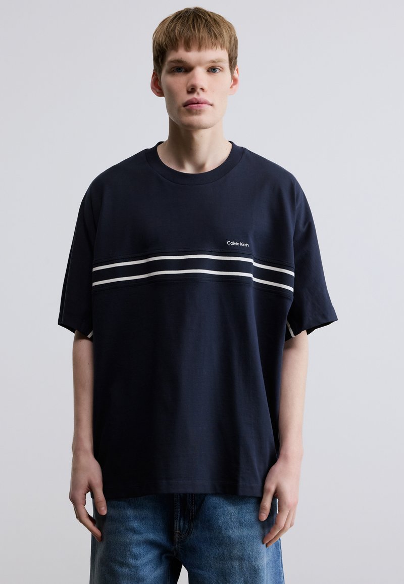 Μπλε ναυτικό βαμβακερό t-shirt με κοντά μανίκια, που διαθέτει μια λευκή οριζόντια ρίγα και ένα μικρό λογότυπο στο στήθος. Χαλαρή εφαρμογή.