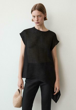 Młoda kobieta o krótkich włosach, ubrana w luźną czarną bluzkę i czarne spodnie, trzymająca małą beżową plecioną torebkę przy gładkiej ścianie.