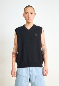 Schwarzer ärmelloser Strickpullover mit V-Ausschnitt, versehen mit einem kleinen Logo. Die Person trägt hellblaue, locker sitzende Hosen.