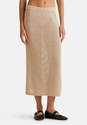 Beige gestrickter Maxikleid mit einem lockeren, offenen Strickmuster und breitem Bund; kombiniert mit schwarzen Netzschuhen.