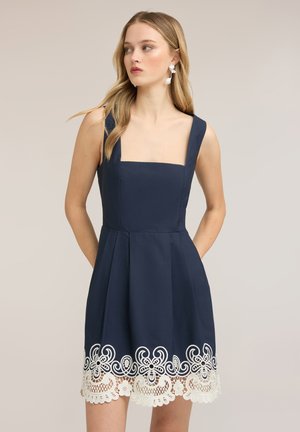Frau mit langen blonden Haaren, die ein ärmelloses marineblaues Kleid mit quadratischem Ausschnitt und weißem Spitzen-Blumenabschluss trägt und zur Seite schaut.