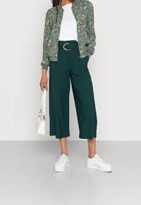 Veste bomber à fleurs vertes, t-shirt blanc, pantalon large sarcelle, baskets blanches et petit sac à main blanc. Tenue décontractée avec une coupe ample.