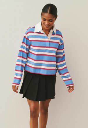 Kobieta w niebiesko-biało-czerwonej bluzce w paski z długim rękawem i białym kołnierzykiem oraz czarnej plisowanej spódnicy, uśmiechająca się i patrząca w dół.