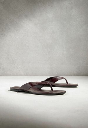 Tumma ruskeat nahkaiset litteät sandaalit, joissa on varvassilmäremmi, esitetty vierekkäin vaalealla pinnalla yksinkertaisella harmaalla taustalla.