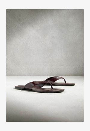 Tumma ruskeat nahkaiset litteät sandaalit, joissa on varvassilmäremmi, esitetty vierekkäin vaalealla pinnalla yksinkertaisella harmaalla taustalla.