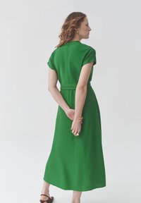 Rochie verde, cu mâneci scurte, cu talie evidențiată printr-un centură, lungime midi, textură netedă și design minimalist, văzută din spate.
