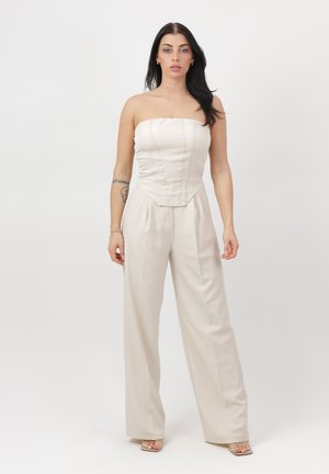 Femme aux longs cheveux noirs portant une combinaison beige bustier avec jambes larges, debout sur fond blanc avec une expression neutre et un tatouage sur le bras gauche.