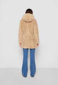 Person iført en beige fuzzy hættejakke og blå flagrende jeans, der står med ansigtet mod en ensfarvet hvid væg, hænderne afslappet langs siden.