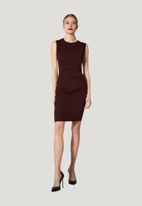 Burgunderfarbenes ärmelloses Kleid mit taillierter Silhouette, Raffung an der Taille und knielangem Saum. Trägt schwarze High Heels.