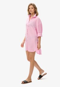 Vestido de camisa de lino rosa con frente de botones, mangas enrolladas y aberturas laterales. Combinado con sandalias negras tipo slide. Acentos de diseño minimalista.