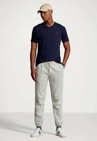 Teget majica s V-izrezom i crvenim logotipom, u kombinaciji s sivim suženim trenirkom. Model nosi bež kapu i bijele tenisice s crnim prugama.