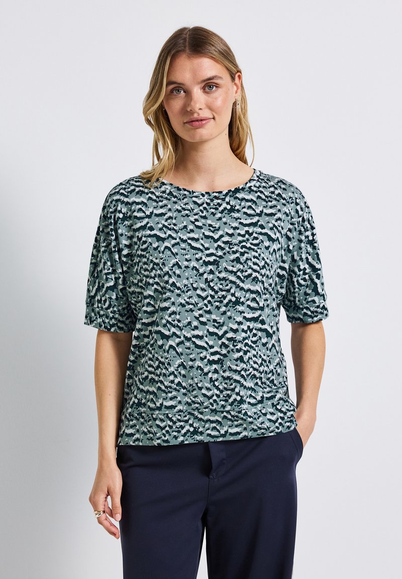 Nainen, joka on pukeutunut sinisen ja mustan sävyistä kuvioituun lyhythihaiseen paitaan, yhdistettynä tummansinisiin housuihin, seisoo yksi käsi taskussa.