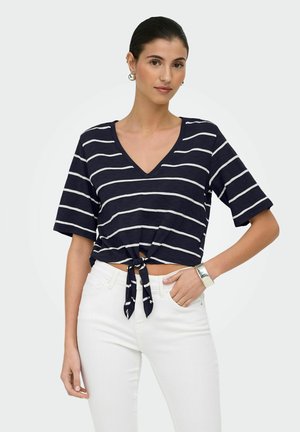 Donna che indossa una maglietta a righe blu navy e bianca con scollo a V e nodo sul davanti, maniche al gomito e pantaloni bianchi, posando con una mano sul fianco.