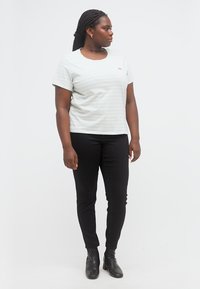 Γυναίκα με πλεξούδες, φοράει λευκό κοντομάνικο πουκάμισο, στενά μαύρα παντελόνια και μαύρα μποτάκια αστραγάλου, στέκεται και κοιτάζει προς τα αριστερά της.