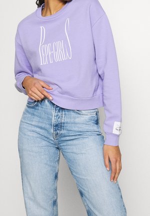 Kobieta w lawendowej bluzie z białym napisem "PEPE-GIRLS" i jasnoniebieskich jeansach z wysokim stanem, ręka spoczywa w pobliżu pasa.