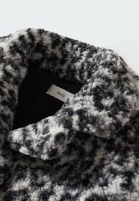 Černobílý texturovaný kabát s knoflíky, vyrobený z měkkého, hebkého materiálu, s širokým límcem a našitou etikou viditelnou na výstřihu.