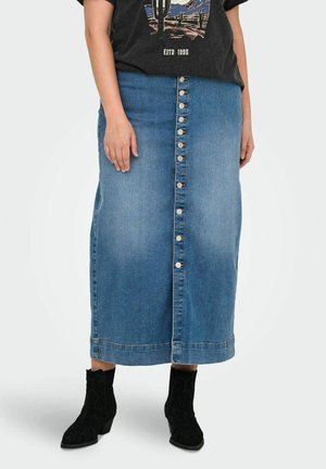 Jupe maxi en denim avec une coupe droite, couleur bleu clair, fermeture à boutons sur le devant et ourlet effiloché. Associée à des bottes noires en daim à la cheville.