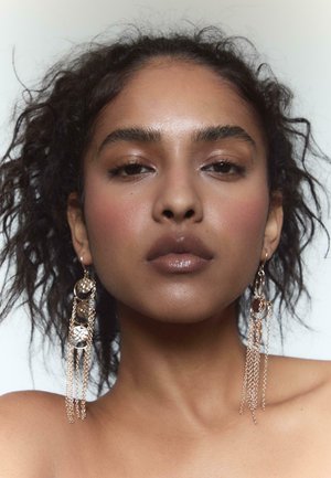 Dlhé visacie náušnice s kruhovými zlatými detailmi a prvkami reťazí, umiestnené na neutrálnom pozadí. Subjekt má kučeravé vlasy.