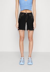 Černé džínové šortky s třepeným lemem, spárované se světle modrým pleteným crop topem a modrými sandály s tvarovaným stélkem a popruhy. Jednoduché pozadí.