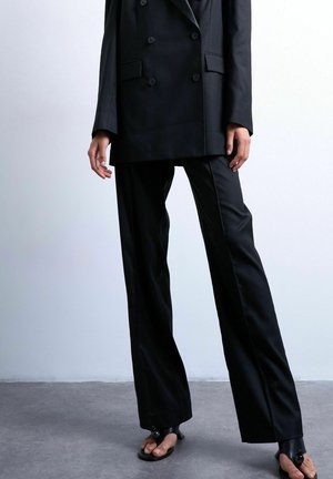 Persoon die een zwart dubbelrijig colbert, wijdvallende zwarte broek en open teen sandalen met hakken draagt, staand op een grijze vloer.