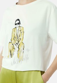 Weißes T-Shirt mit kurzen Ärmeln, das eine gedruckte Illustration einer Figur in Gelb zeigt, die Sonnenbrillen trägt, mit dem Text "Ermächtigt und Unaufhaltbar."