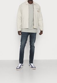 Kremowa kurtka bomberka na szarym swetrze, zestawiona z ciemnymi dżinsami i granatowymi trampkami. Kurtka ma kieszeń z naszywką z logo.