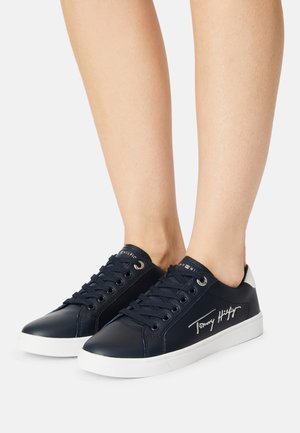 Marineblauwe lage sneakers met witte zolen en het "Tommy Hilfiger"-logo, gedragen aan de voeten tegen een witte achtergrond.