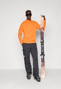 Orangene Zip-up Langarmshirt, dunkle Cargo-Hosen, schwarze Sonnenbrille, hält einen Ski mit braunen und schwarzen Akzenten. Schneestiefel runden das Outfit ab.