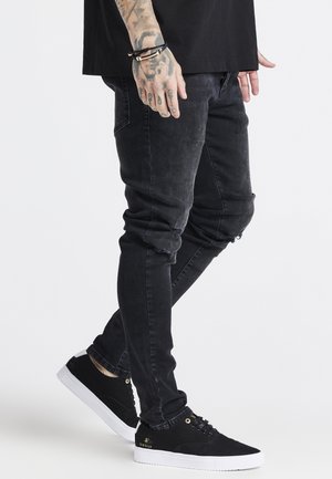 Person iført sorte revnede skinny jeans, sorte sneakers med hvide såler og et sort armbånd på den tatoverede venstre hånd.