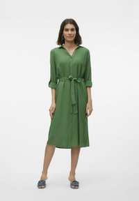 Abito verde con bottoni e cintura, maniche lunghe arrotolate e tessuto liscio. Indossato con ciabatte blu.