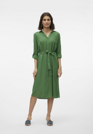 Abito verde con bottoni e cintura, maniche lunghe arrotolate e tessuto liscio. Indossato con ciabatte blu.