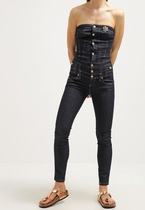 Mørk denim jumpsuit med stropløs design, dekorative knapper foran, tætsiddende pasform og lommedetaljer; båret med brune sandaler.