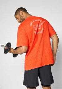 Rød bomulds T-shirt med rund hals, prydet med et hvidt grafisk print på ryggen. Modellen holder en sort håndvægt og har sorte shorts på.
