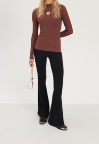 Femme portant un haut à manches longues côtelé marron, un pantalon évasé noir, des chaussures noires à bouts métalliques, et tenant un petit sac à main blanc avec une chaîne dorée.