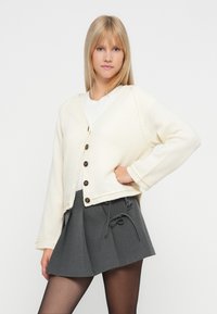 Cremefarbener gestrickter Cardigan mit V-Ausschnitt, großen braunen Knöpfen und gerippten Bündchen, kombiniert mit einem grauen Faltenrock und schwarzen Strumpfhosen.