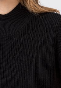 Svart stickad tröja med hög ribbad halsringning och textilstruktur, som framhäver vertikala mönster över hela plagget.