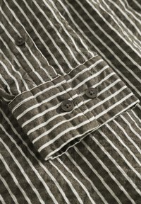 Striped button-up shirt featuring dark olive green fabric with white stripes, two brown buttons on the cuff, and a textured weave pattern.