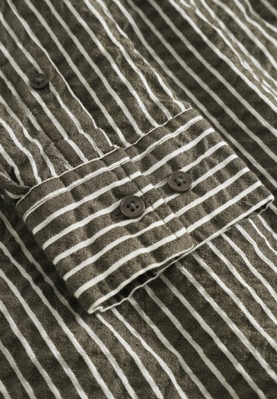 Chemise à rayures avec un tissu vert olive foncé et des rayures blanches, deux boutons marron sur le poignet et un motif de tissage texturé.