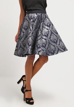 Femme portant une jupe mi-longue bleu marine et argent à motifs, des sandales noires à plateforme et talons hauts, et un haut noir sans manches.