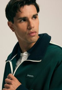 Zielona kurtka z zamkiem błyskawicznym i żebrowanym granatowym kołnierzem, białym zamkiem błyskawicznym oraz małą naszywką z logo. Noszona na białej koszuli z kołnierzykiem.