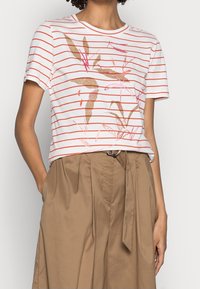Camiseta de manga corta blanca a rayas con estampado botánico en rosa y dorado, combinada con pantalones de talle alto beige con un cinturón atado.
