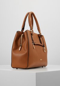 Sac à main en cuir marron texturé avec deux poignées, fermetures éclair dorées et un pendentif circulaire en or, posé sur un piédestal blanc sur un fond uni.