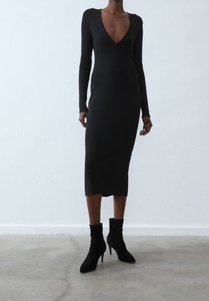 Vestido midi negro con canalé, de mangas largas y escote en V pronunciado. Combinado con botas de ante negras hasta el tobillo con tacón de aguja.
