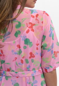 Blommig kimono i semi-genomskinligt rosa tyg med flerfärgade abstrakta mönster; har ett bälte och lösa ärmar för en avslappnad passform.