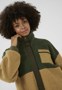 Pullover in pile verde e beige, con tasca sul petto con zip, colletto alto e polsini a coste. Tessuto texturizzato con una finitura morbida.