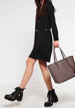 Femme en robe noire avec ceinture marchant, portant des bottes noires à lacets, et transportant un grand sac cabas taupe avec deux poignées.