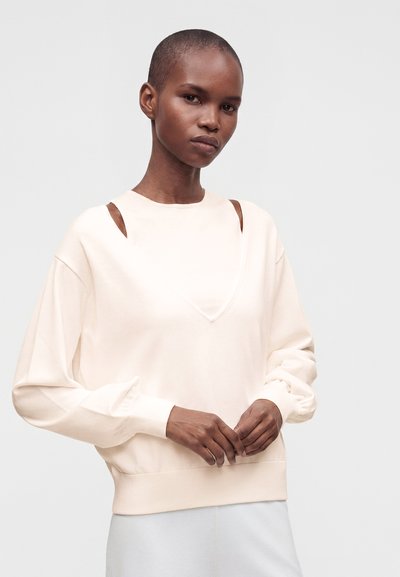 Femme aux cheveux courts portant un haut ample à manches longues blanc cassé avec des découpes aux épaules, debout devant un fond uni.