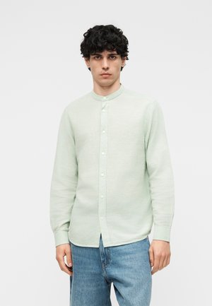 Joven con cabello rizado y oscuro, vistiendo una camisa de botones color verde pálido con cuello mandarín y jeans azul claro, de pie frente a un fondo liso.