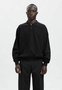 Czarna koszulka polo z długim rękawem, wykonana z miękkiego materiału o luźnym kroju, z trzyguździową plisą i ściągaczami na mankietach, zestawiona z ciemnymi spodniami.