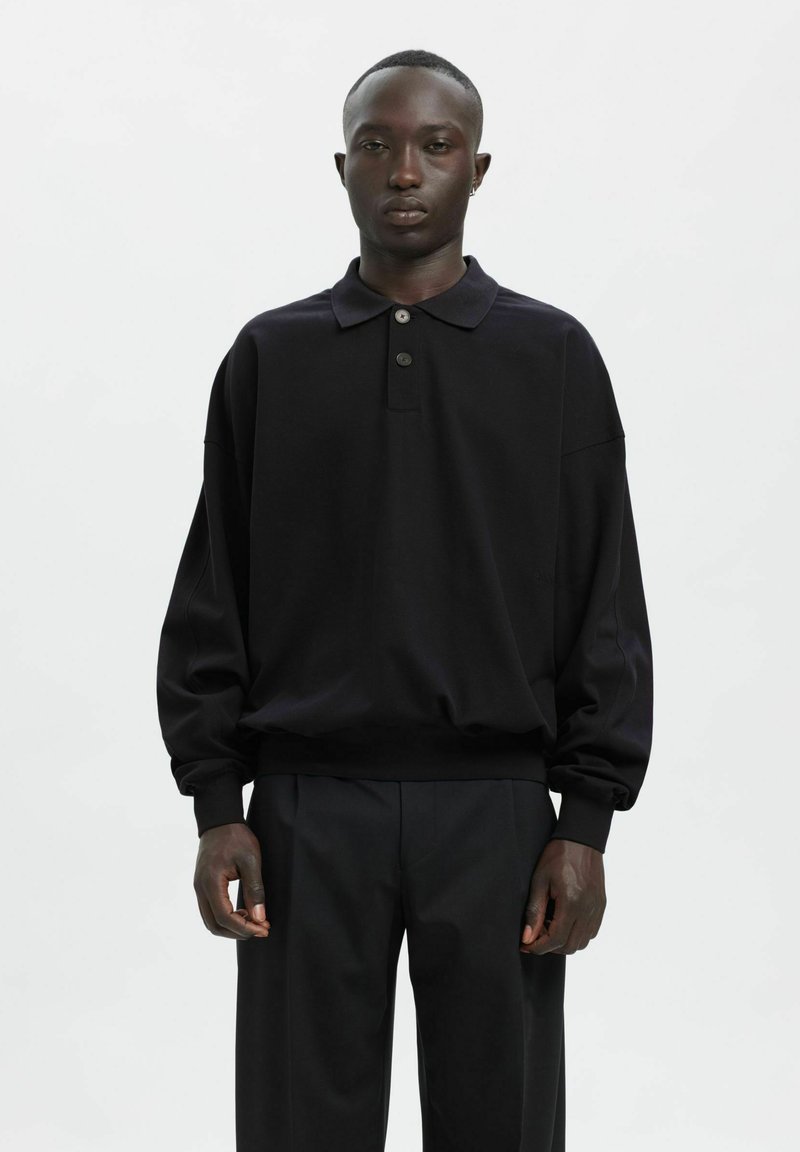 Czarna koszulka polo z długim rękawem, wykonana z miękkiego materiału o luźnym kroju, z trzyguździową plisą i ściągaczami na mankietach, zestawiona z ciemnymi spodniami.